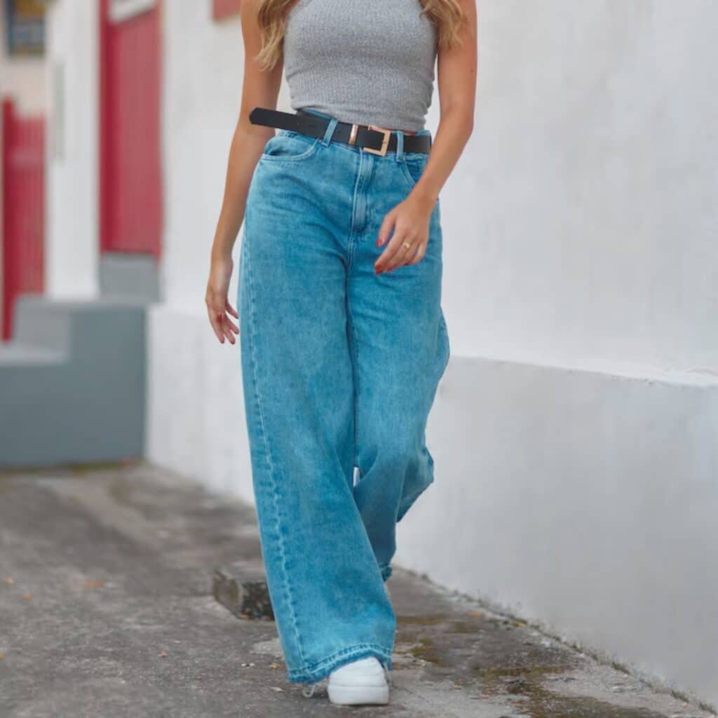 A stylish woman walks outdoors along a white wall with red-trimmed windows, wearing a gray sleeveless ribbed top, high-waisted wide-leg jeans, a black belt, and white sneakers.