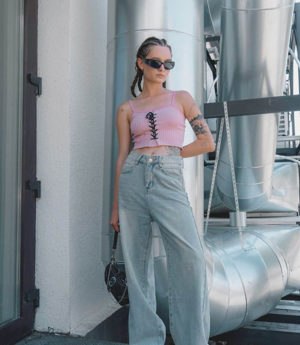A young woman stands confidently outdoors near industrial metal ducts. She wears a pink ribbed crop top with black lace-up detail, high-waisted light-wash jeans, black sunglasses, and small braided hairstyle. She holds a small black purse with a chain strap and poses against a backdrop of metallic piping.