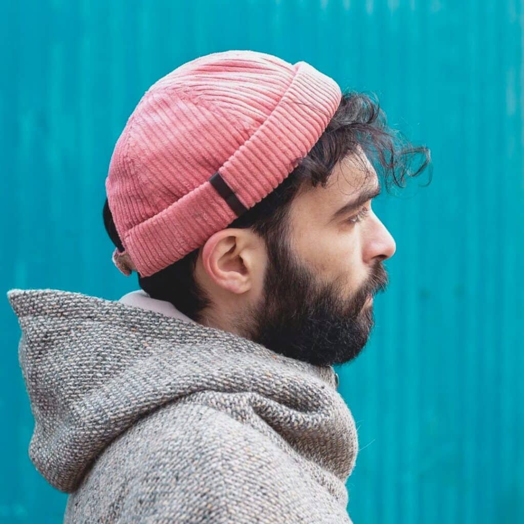 A profile view of a bearded man wearing a pink corduroy beanie and a speckled gray hooded jacket, standing in front of a bright turquoise corrugated metal wall.