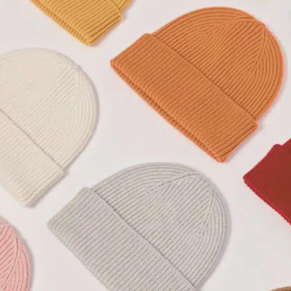 "Flat lay of multiple knit beanies in colors like yellow, orange, white, gray, pink, and red on a white background, arranged in a scattered, balanced pattern with ribbed textures.